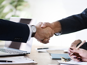 Two professionals shake hands across a desk, with a laptop and documents nearby, signaling a business agreement.