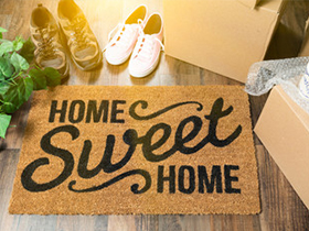 Doormat reading 'Home Sweet Home' on a wooden floor, with shoes, a plant, and moving boxes nearby.
