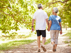 Two older adults walking hand in hand along a sunlit park path.